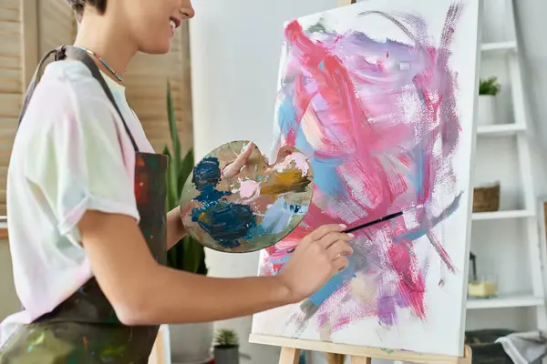 Una joven con el pelo corto crea alegremente una pintura abstracta en su acogedor espacio de estudio en casa. - foto de stock