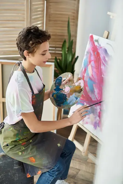 A young woman with short hair enjoys painting on a canvas at home, expressing her artistic passion. — Stock Photo
