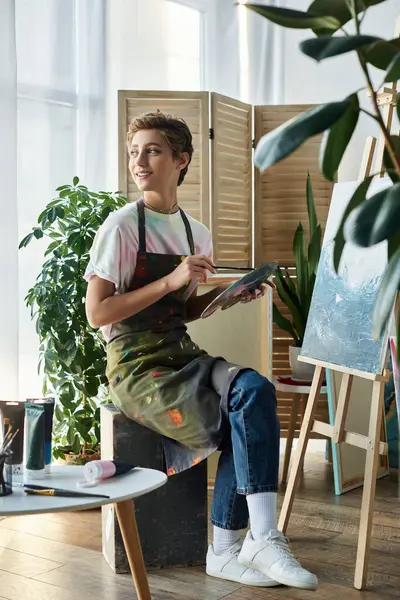 A young woman with a relaxed demeanor paints on a canvas in her cozy home studio surrounded by greenery. — Stock Photo