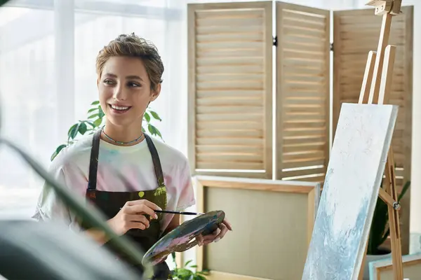 Inside a bright room, a young woman with short hair joyfully paints a colorful picture while smiling. — Stock Photo