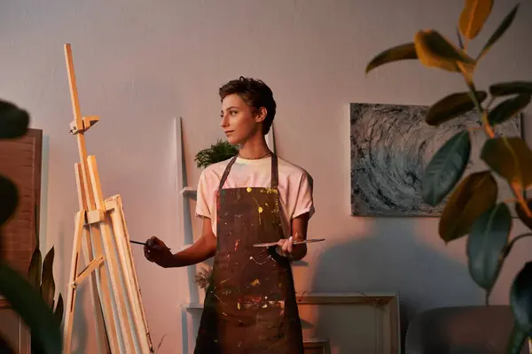 A young woman with short hair explores her artistic side by painting at home, surrounded by nature and warmth. — Stock Photo