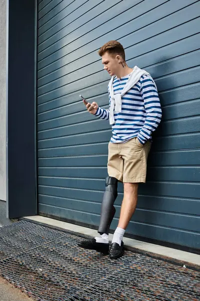 A stylish young man leans against a blue wall, engaging with his smartphone while wearing shorts. — Stock Photo