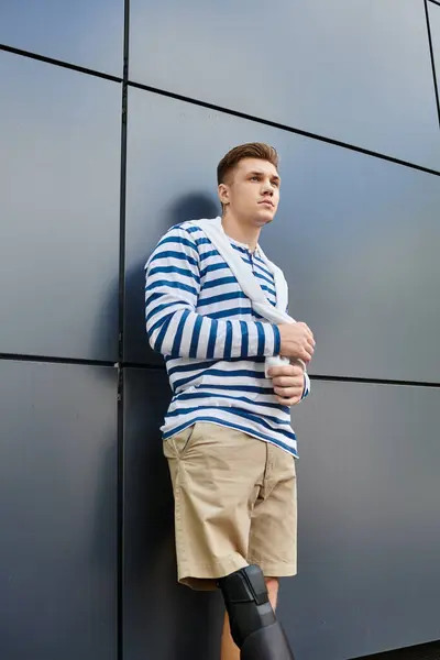 Un joven está de pie pensativamente con una pierna protésica, vestido casualmente en rayas por una pared elegante. — Stock Photo