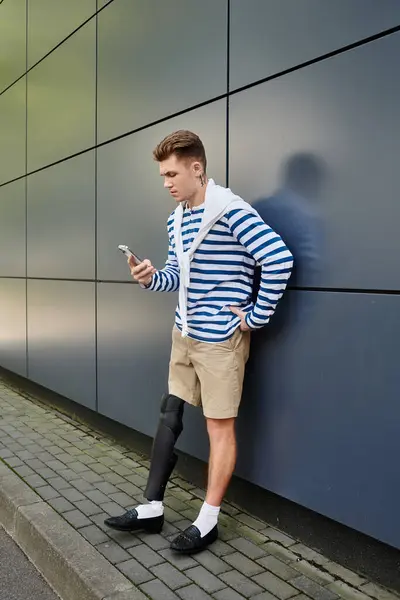 A stylish young man stands against a sleek wall, absorbed in his phone, showcasing resilience. — Stock Photo