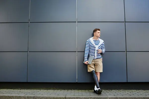 A young handsome man leans against a sleek wall, showcasing his stylish attire and prosthetic leg. — Stock Photo