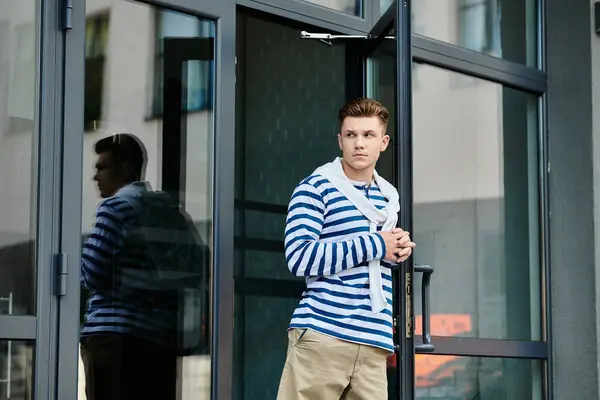 Un joven vistiendo un suéter a rayas y caqui mira pensativamente fuera de una puerta de cristal. - foto de stock