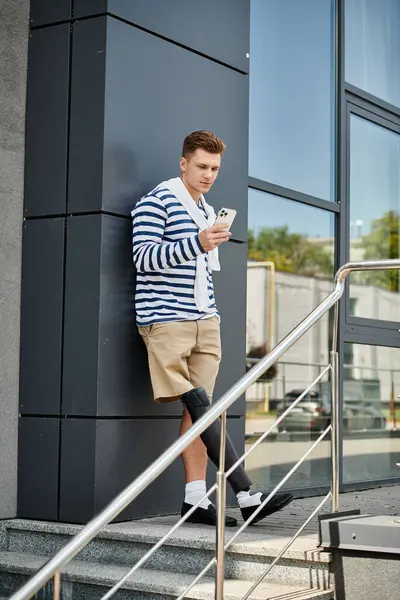 A young, stylish man leans against a modern building, focused on his smartphone. — Stock Photo