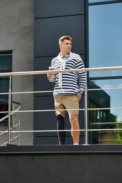 A young man confidently holds a coffee on a balcony, proudly showing his prosthetic leg. — Stock Photo