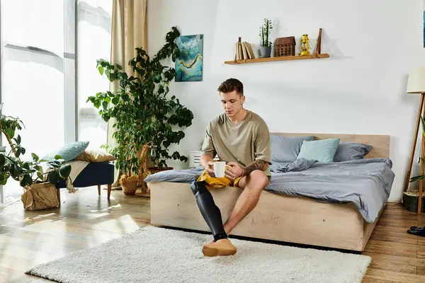 A stylish young man sits on his bed, sipping coffee while appreciating his serene surroundings. — Stock Photo