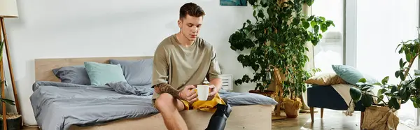 A young man with a prosthetic leg sits on a bed, reflecting in a sunlit, plant filled room. — Stock Photo