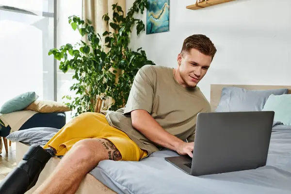 Un giovane uomo gode di lavorare sul suo computer portatile mentre seduto su un letto in una stanza elegante. — Foto stock