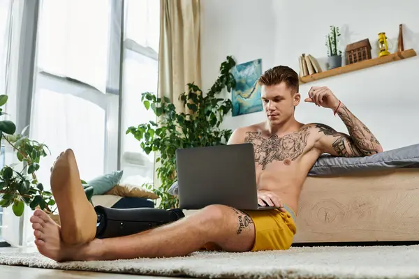 A young man with tattoos sits on a rug, focused on his laptop while showing his prosthetic leg. — Stock Photo