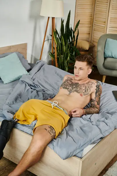 In una camera da letto tranquilla, un bel giovane uomo si rilassa senza maglietta, mostrando tatuaggi e una gamba protesica. — Foto stock