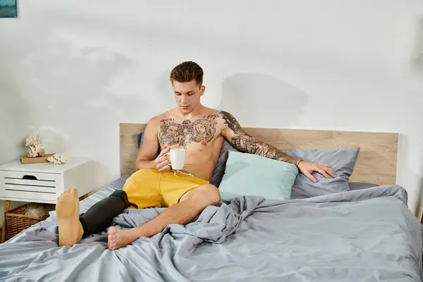 Este joven, con un tatuaje detallado, se sienta cómodamente en su cama, disfrutando de una bebida caliente. — Stock Photo