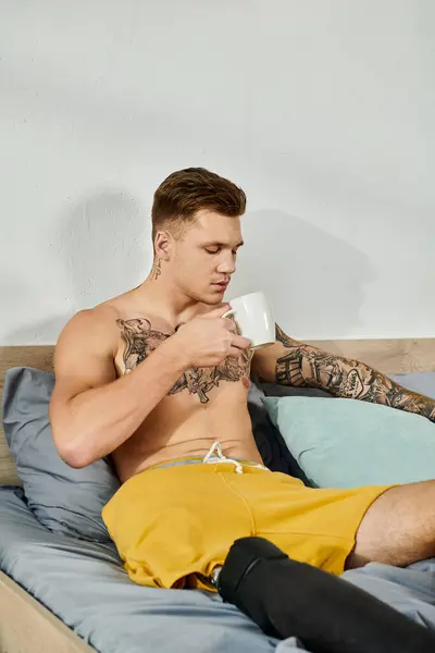 A handsome young man with tattoos relaxes on his bed, sipping coffee with a prosthetic leg. — Stock Photo