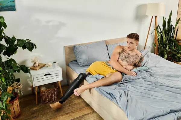 Un joven guapo se sienta casualmente en una cama, mostrando su pierna protésica y tatuajes. - foto de stock