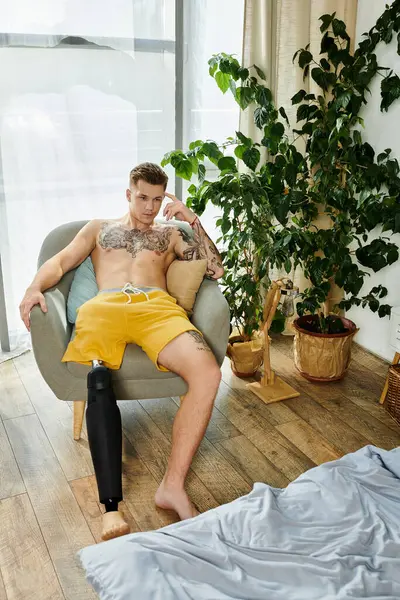 A young man sits comfortably in an armchair, showcasing his prosthetic leg and tattoos. — Stock Photo
