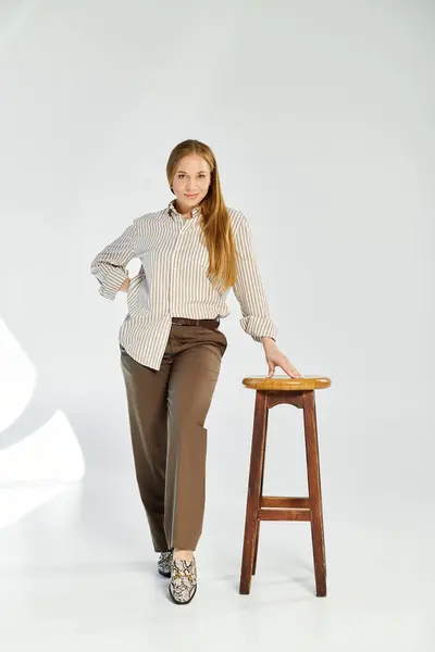 A woman with long hair in a striped shirt poses next to a wooden stool against a neutral grey background. — Stock Photo