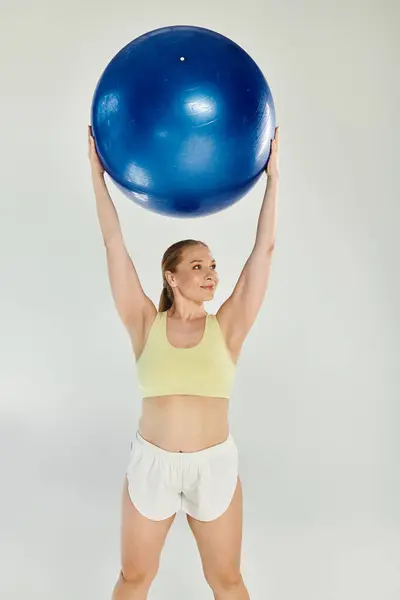 A fit woman joyfully lifts a large blue exercise ball in a bright studio space, showcasing her strength. — Stock Photo