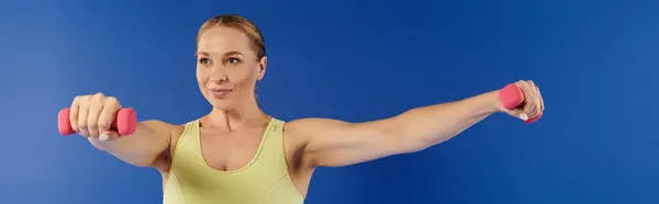 A dedicated sportswoman engages in a workout routine, using pink dumbbells in a studio with blue background. — Stock Photo