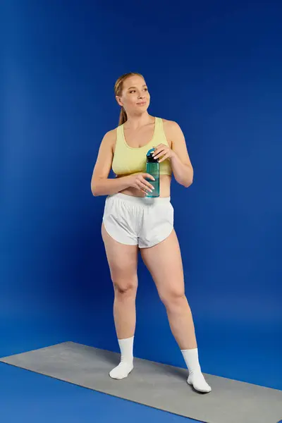 A fit woman wearing a crop top pauses with a water bottle, reflecting her dedication in a bright studio. — Stock Photo