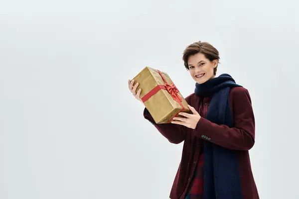 A happy woman in stylish winter wear smiles with a gold gift box tied with a red ribbon. — Stock Photo