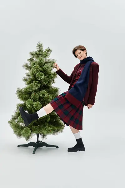 A fashionably dressed woman playfully interacts with a decorative winter tree indoors. — Stock Photo
