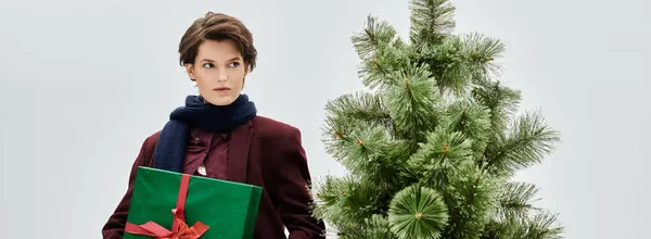 A stylish woman in winter wear stands by a decorated Christmas tree, holding a gift. — Stock Photo