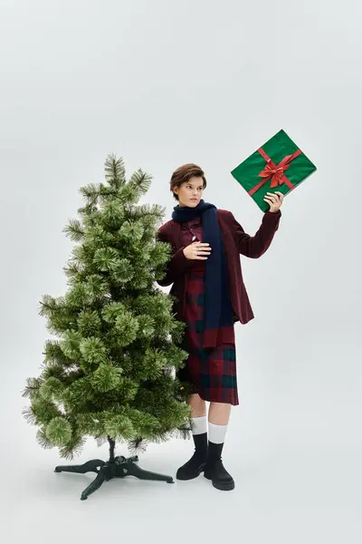 A lovely woman in cozy winter fashion holds a gift by the Christmas tree. — Stock Photo