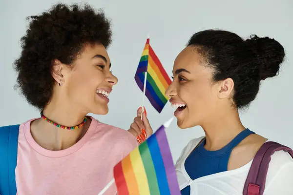 Two cheerful friends smile at each other while waving rainbow flags, celebrating their bond. — Stock Photo