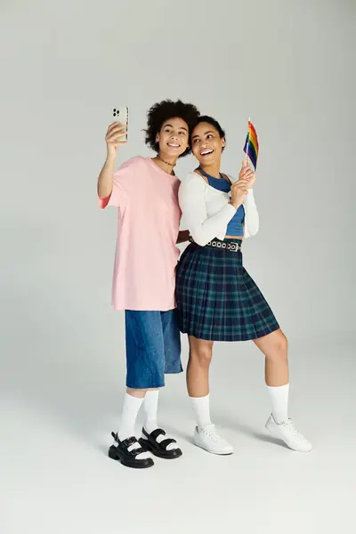 Two friends happily embrace their unique styles while holding a rainbow flag and taking a selfie. — Stock Photo