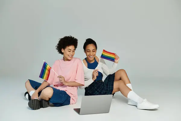 Two young women sit on the floor, waving rainbow flags and enjoying a celebratory moment together. — Stock Photo