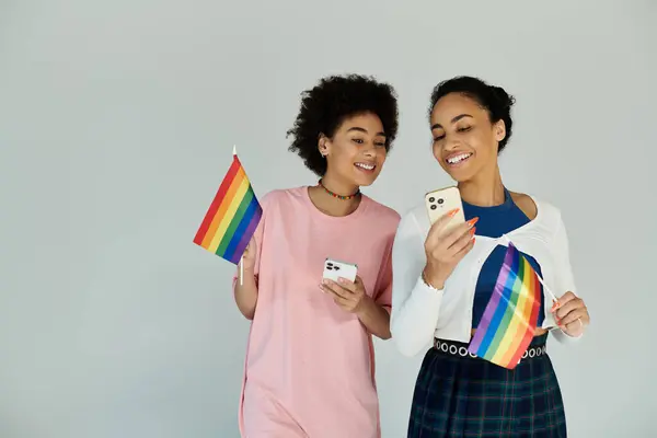 Two young women proudly hold rainbow flags while looking at their phones with joy. — Stock Photo