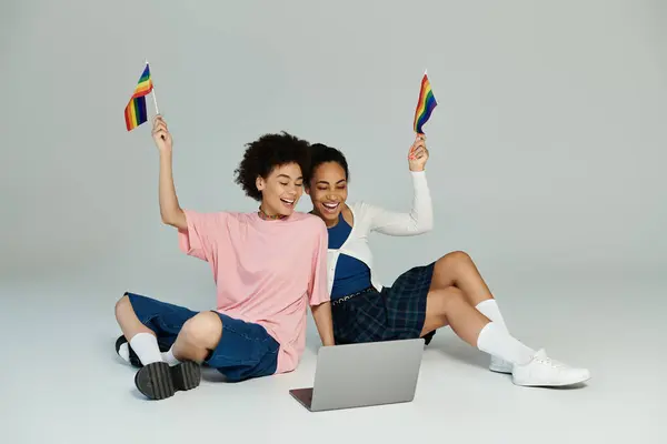 Two friends express excitement while engaging with a computer, waving colorful flags joyfully. — Stock Photo