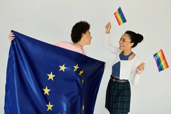 Two joyful friends embrace diversity, holding the EU flag and rainbow flags in celebration. — Stock Photo