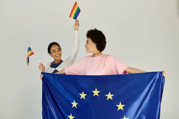 Two friends joyfully wave rainbow flags while holding an EU flag, symbolizing pride and inclusivity. — Stock Photo