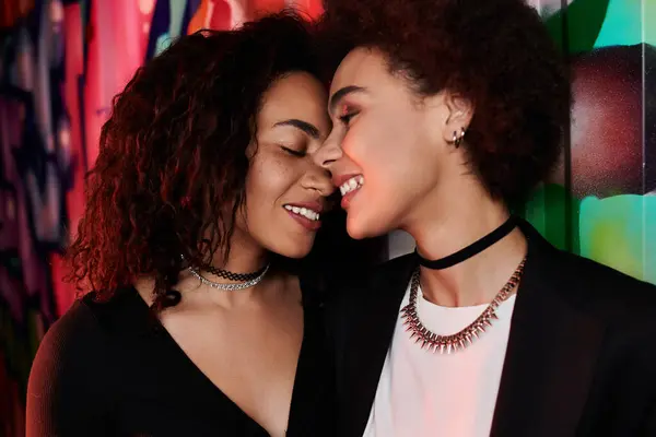 Two women lean close together, smiling affectionately against a colorful wall in the evening. — Stock Photo