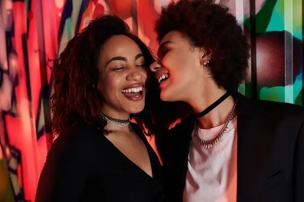 Two friends share laughter and joy, illuminated by colorful lights and vivid artwork behind them. — Stock Photo