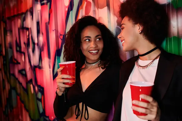 Two women share joyful moments with drinks in hand amidst a striking graffiti backdrop. — Stock Photo