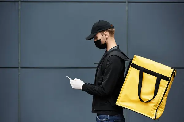 Um homem de entrega usando uma máscara preta e luvas verifica os detalhes da ordem enquanto está em pé perto de um prédio de escritórios. — Fotografia de Stock