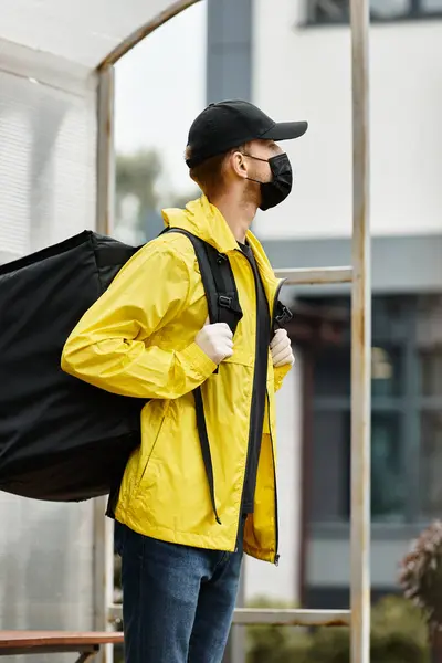 A delivery man wearing a yellow jacket and black mask enters a contemporary office building carrying an order. — Stock Photo