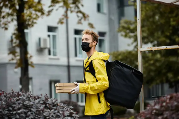 Un fattorino che indossa una maschera medica nera trasporta pacchi in un edificio per uffici in un giorno di pioggia. — Foto stock