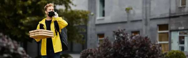 Un livreur portant un masque noir transporte plusieurs boîtes à pizza en parlant au téléphone devant un bureau. — Photo de stock