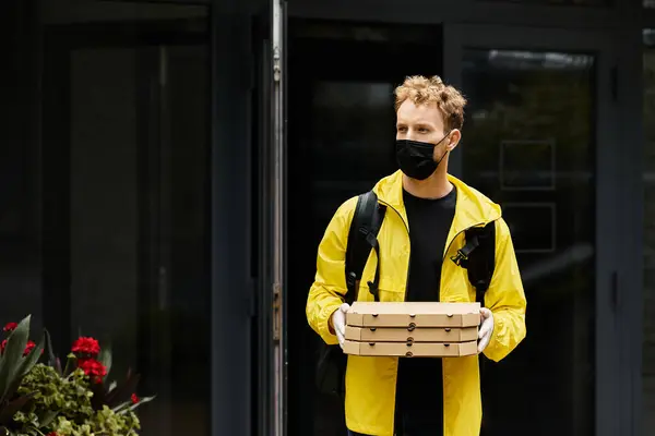 Una persona di consegna che indossa una maschera medica nera trasporta più scatole di pizza in un edificio per uffici. — Foto stock