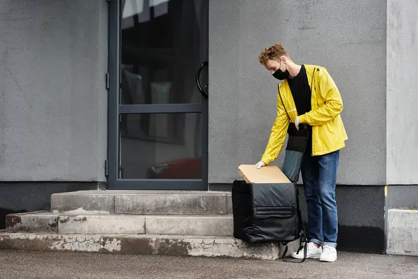 Um homem de entrega em uma máscara preta traz um pacote para a entrada de um prédio de escritórios em um dia ensolarado. — Fotografia de Stock