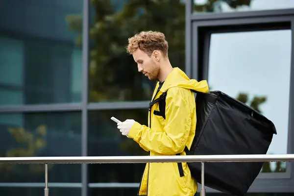 A delivery man in a yellow rain jacket checks his phone while bringing an order into an office building. — Stock Photo