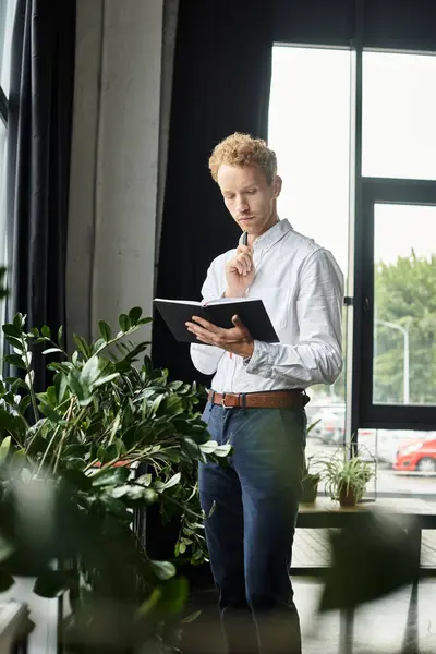 A focused redhead in a white shirt analyzes his project on a tablet in a stylish office surrounded by greenery. — Stock Photo