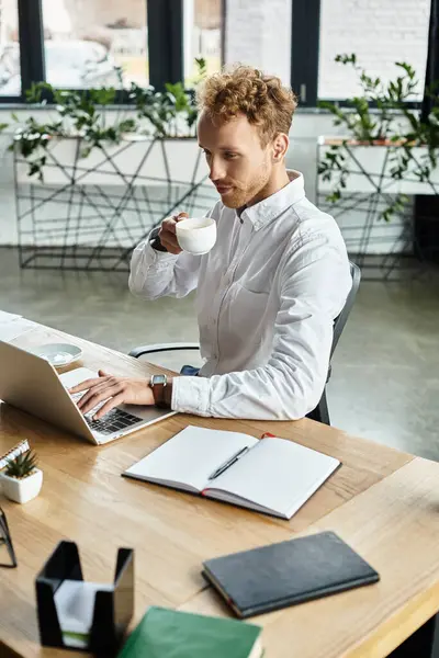 Un uomo d'affari rossa con una camicia bianca lavora sul suo computer portatile, sorseggiando caffè in uno spazio ufficio contemporaneo. — Stock Photo