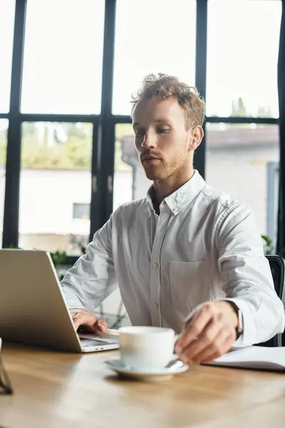 Una rossa con una camicia bianca si concentra sul suo computer portatile mentre si gode il caffè in uno spazio di lavoro contemporaneo. — Foto stock