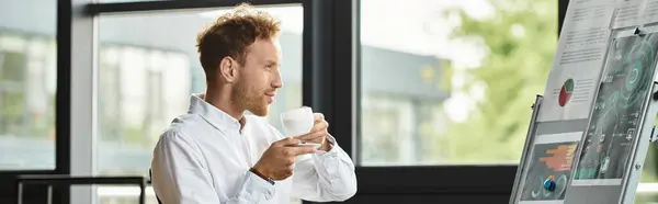 Un hombre de negocios pelirrojo con una camisa blanca bebe café mientras revisa intensamente los detalles del proyecto en una pizarra. - foto de stock